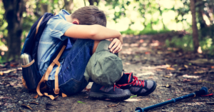 Boy sitting on the ground with a backpack on covering his face with his arms