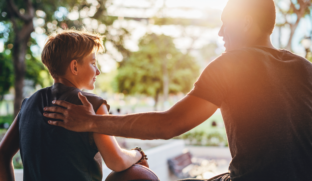 A son sitting next to his dad outside. The son has his arm leaning on a basketball and the dad has his left hand on his son's shoulder in a reassuring way.