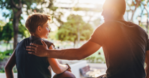 A son sitting next to his dad outside. The son has his arm leaning on a basketball and the dad has his left hand on his son's shoulder in a reassuring way.